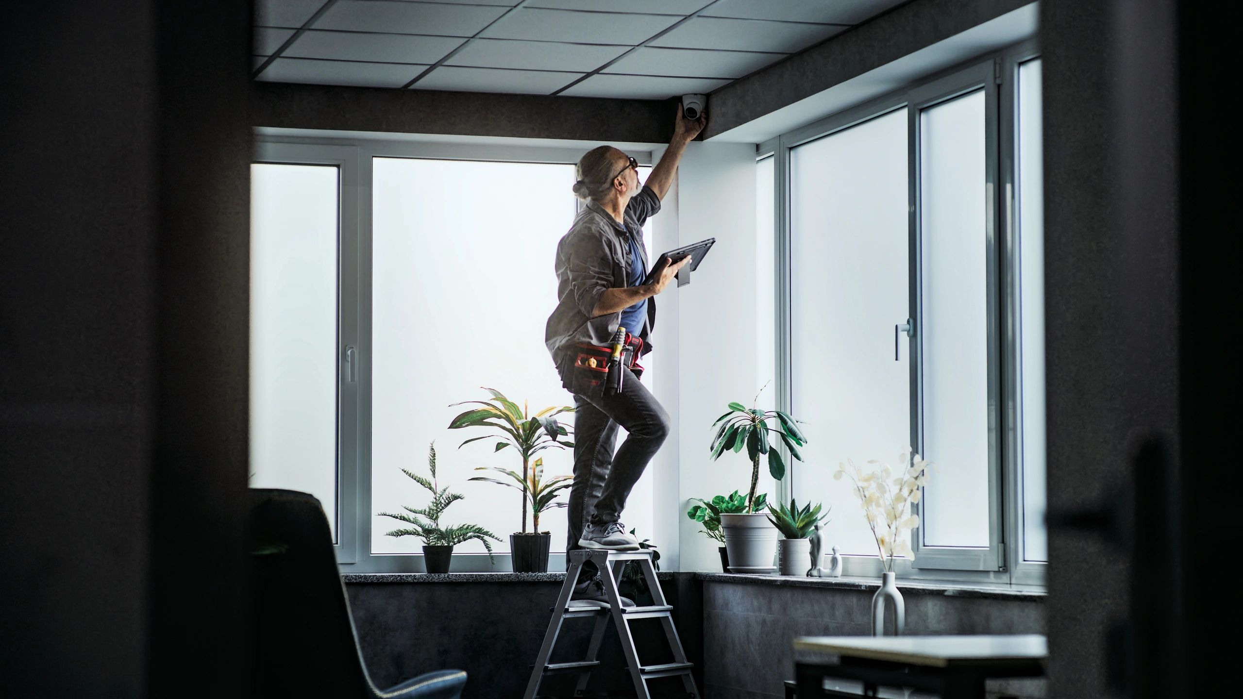 Male technician stands on ladder, installs CCTV camera in office room using tablet